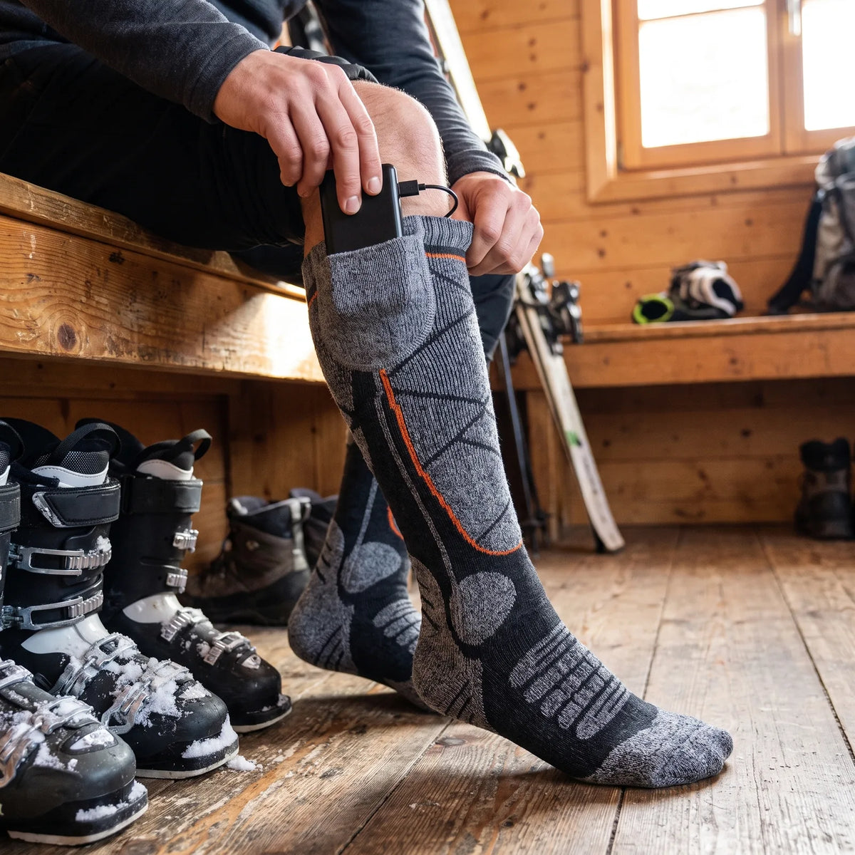 Akku wird in die Tasche am Bund der beheizten Socke gesteckt, Skischuhe am Boden